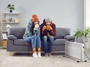 Young Frozen Couple Sitting At Home In Winter Outerwear And Trying To Keep Warm.