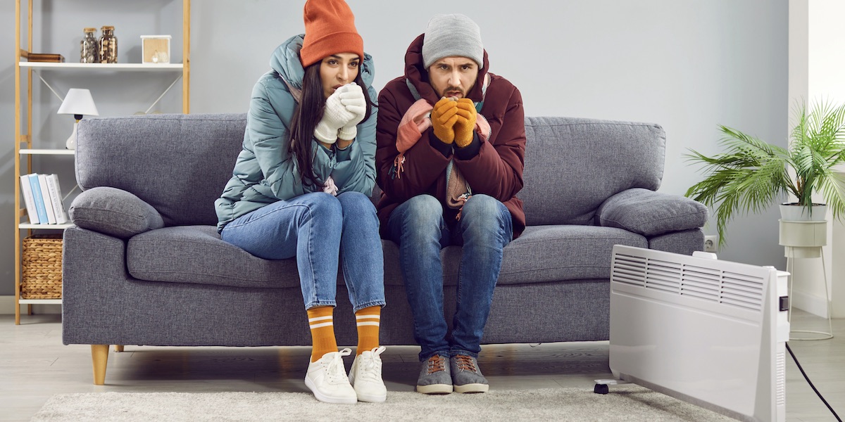 Young Frozen Couple Sitting At Home In Winter Outerwear And Trying To Keep Warm.